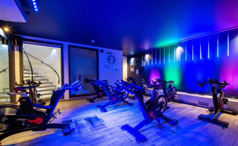 A modern spinning class room with several stationary bikes arranged in rows. The room features vibrant, colorful lighting on the walls and ceiling, creating an energetic atmosphere. The wooden floor and contemporary design add a stylish touch to the fitness space.