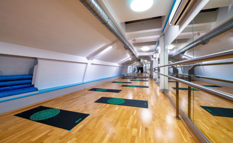 A modern, well-lit yoga studio with mirrors along one wall, wooden flooring, and several colorful yoga mats evenly spaced on the floor. Blue rolled-up exercise mats are neatly stored in a nook. A ballet barre runs along the mirrored wall.