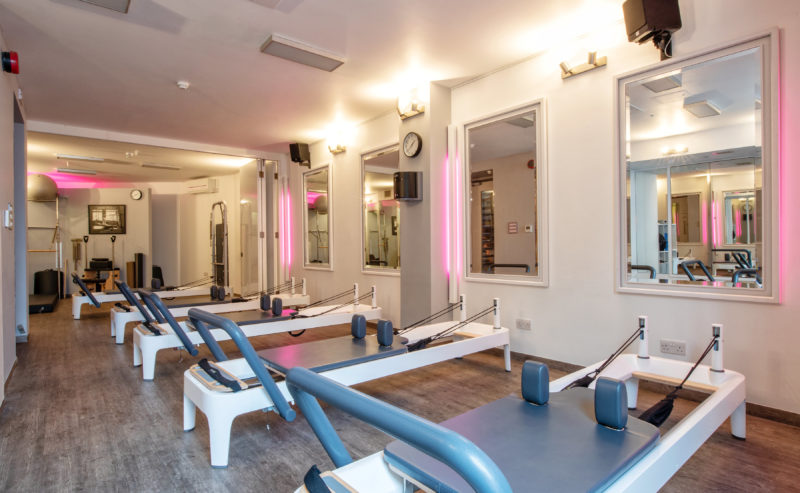 A spacious, well-lit Pilates studio with several reformer machines lined up. The room features large mirrors on the walls, a wooden floor, and a clean, modern design. Some equipment is neatly stored in the background, and the lighting creates a relaxed atmosphere.